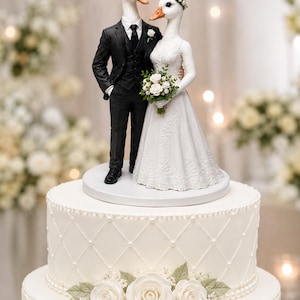 May include: A wedding cake topper featuring two anthropomorphic geese standing on a white circular base. One goose wears a black three-piece suit with a white shirt and black tie. The other goose wears a white lace gown and a floral crown.