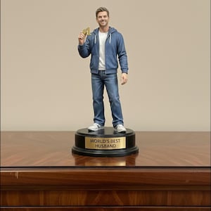 May include: A figurine of a man wearing a blue hoodie, jeans, and white sneakers, holding a gold object. The figurine stands on a black base with a gold plaque that reads "WORLD'S BEST HUSBAND".
