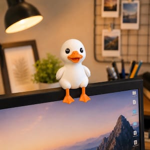 Könnte beinhalten: Eine weiße Entenfigur mit glatter Oberfläche, ausgestattet mit einem leuchtend orangefarbenen Schnabel und orangefarbenen Schwimmfüßen. Die Ente hat große schwarze Augen und ist so gestaltet, dass sie auf dem oberen Rand eines Monitors sitzt.