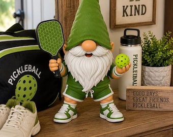 May include: A garden gnome figurine wearing a green hat, green shirt, and green shorts, holding a green pickleball paddle in one hand and a yellow pickleball in the other. The gnome wears white sneakers with green accents and white wristbands.