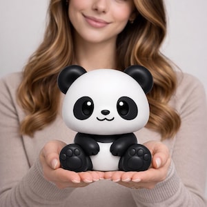 Może przedstawiać: Biało-czarna figurka pandy z dużymi oczami, czarnymi uszami i łapami. Panda trzymana jest w dłoniach osoby. Figurka ma uśmiechniętą minę i jest uroczym, kreskówkowym projektem.