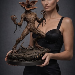 Może przedstawiać: Szczegółowa, brązowa statua Demogorgona z serialu Stranger Things. Postać potwora ma głowę w kształcie kwiatu, ostre pazury i stoi na skalistej podstawie. Statua jest trzymana przez osobę w czarnej sukience.