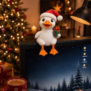 Könnte beinhalten: Eine weiße Entenfigur mit orangefarbenen Füßen und Schnabel, die eine rote Weihnachtsmütze mit weißem Rand trägt. Die Ente hält ein kleines grünes Geschenk mit roter Schleife und sitzt auf der Oberkante eines Computerbildschirms.