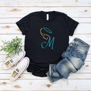 Peut inclure: T-shirt noir avec un motif turquoise et or représentant le profil d'une femme, une auréole et la lettre "M". Le t-shirt est associé à un jean bleu et à des baskets en toile blanches. Les baskets ont des détails rouges et le mot "converse".