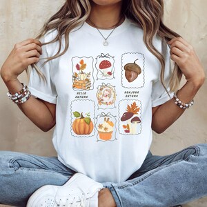 Peut inclure: T-shirt blanc orné d'illustrations sur le thème de l'automne. Les motifs incluent des citrouilles, des champignons, des glands, avec les textes "Hello Autumn" et "Bonjour Autumn". Un vêtement décontracté et saisonnier.