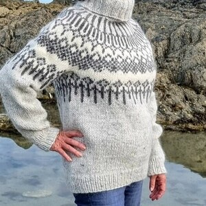 Puede incluir: Un su&eacute;ter de punto blanco y gris con un patr&oacute;n tradicional escandinavo. El su&eacute;ter tiene cuello alto y mangas largas.
