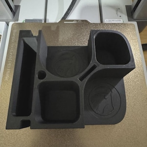 May include: A matte black 3D printed desk organiser featuring multiple compartments of varying shapes and sizes. The organiser includes a long rectangular slot, a square bin, a deep rounded cup, and two circular recessed areas embossed with a horse head logo.