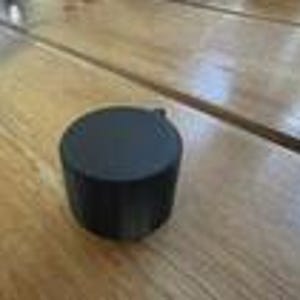 May include: A small, cylindrical black container with a flat, circular lid. The object features a matte finish and a smooth, solid exterior surface. It is a compact, portable item designed with a simple, uniform shape and a neutral dark colour.