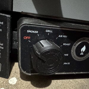 May include: Close-up of a black appliance control panel with a dial and buttons. The dial has settings for "Smoker", "Grill", "Air Fry", "Roast", and "4KE". The buttons are labelled "OFF" and "DRATE". The panel also features a logo that says "Woodfire Flavour Technology".