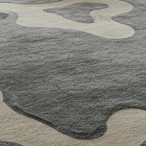 K&ouml;nnte beinhalten: Nahaufnahme eines grauen und beigen Shaggy-Teppichs mit modernem, abstraktem Design. Der Teppich hat eine weiche, pl&uuml;schige Textur.