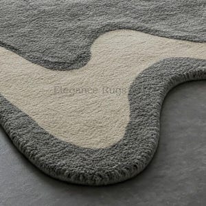 K&ouml;nnte beinhalten: Nahaufnahme eines grauen und cremefarbenen Teppichs mit einem einzigartigen, abstrakten Design. Der Teppich hat eine wellenf&ouml;rmige, organische Form und eine weiche, pl&uuml;schige Textur.