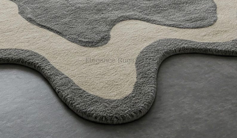 K&ouml;nnte beinhalten: Nahaufnahme eines grauen und cremefarbenen Teppichs mit einem einzigartigen, abstrakten Design. Der Teppich hat eine wellenf&ouml;rmige, organische Form und eine weiche, pl&uuml;schige Textur.
