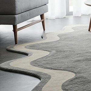 K&ouml;nnte beinhalten: Ein grauer und wei&szlig;er, wellenf&ouml;rmiger Teppich mit modernem Design. Der Teppich hat einen hellgrauen Hintergrund mit einer wei&szlig;en, wellenf&ouml;rmigen Linie, die durch die Mitte verl&auml;uft. Der Teppich ist aus einem weichen, pl&uuml;schigen Material gefertigt und perfekt, um jedem Raum einen Hauch von modernem Stil zu verleihen.