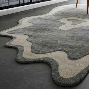 K&ouml;nnte beinhalten: Ein grauer und cremefarbener Teppich mit einem wellenf&ouml;rmigen, abstrakten Design. Der Teppich hat eine weiche, pl&uuml;schige Textur und ist aus hochwertigen Materialien gefertigt. Der Teppich ist perfekt, um jedem Raum einen Hauch von modernem Stil zu verleihen.