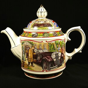 Puede incluir: Una tetera de porcelana blanca con ribetes dorados y un diseño colorido que presenta un coche antiguo, personas y un edificio. La tetera tiene una tapa con un pomo dorado.