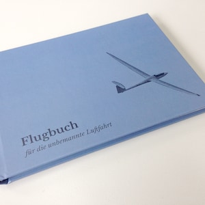 Könnte beinhalten: Ein hellblaues Hardcover-Flugbuch mit einer dunkelblauen Grafik eines Segelflugzeugs auf dem Einband. Der Text auf dem Einband lautet "Flugbuch" in fetter Schrift, gefolgt von dem kleineren Text "für die unbemannte Luftfahrt" in Standardschrift.