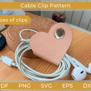 Może przedstawiać: Różowy klips do kabli w kształcie serca ze srebrnym zatrzaskiem, trzymający białe przewody i słuchawki. Na zdjęciu widoczny jest również granatowy klips, korek i tekst "Cable Clip Pattern" oraz "6 types of clips."