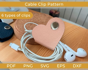May include: A heart-shaped, blush pink cable clip with a silver snap, holding white cords and earbuds. The image also shows a dark blue clip, a cork coaster, and the text "Cable Clip Pattern" and "6 types of clips."