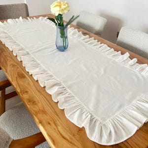 May include: A white rectangular table runner with ruffled edges is displayed on a wooden table. A blue vase with a yellow flower and greenery sits in the center. The table is surrounded by chairs.
