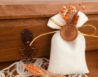 May include: A small white fabric pouch with a gold ribbon and a brown wax seal. The pouch is filled with dried flowers and wheat. The pouch is sitting on a white crocheted doily.