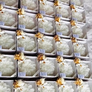 Puede incluir: Una colección de velas blancas con forma de flor en pequeñas cajas blancas con una cinta dorada y flores secas. Cada caja tiene una etiqueta blanca con el texto "Rater & Darina" y una fecha.
