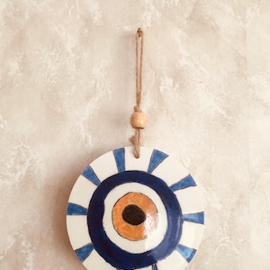 May include: A white ceramic wall hanging with a blue and orange evil eye design. The hanging has a jute rope with a wooden bead.