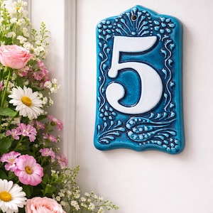 Peut inclure: Panneau de numéro de maison en céramique turquoise avec le chiffre cinq blanc et une bordure florale décorative. Le panneau est rectangulaire avec un sommet incurvé. Un bouquet de roses roses, de marguerites et d'autres fleurs est dans le coin.