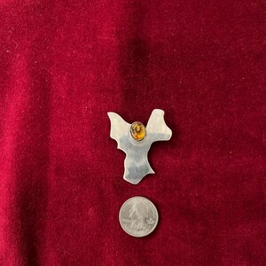 May include: A silver-toned metal brooch with an abstract design, featuring a central amber-colored stone. A U.S. quarter coin is placed below the brooch for size comparison. The brooch is set against a deep red velvet background.