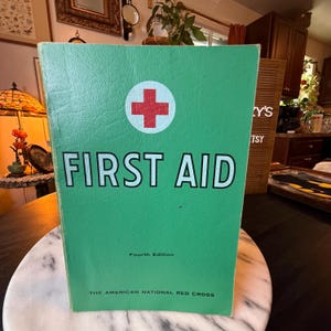 Puede incluir: Un libro de primeros auxilios verde, cuarta edición, de The American National Red Cross. La portada presenta un círculo blanco con una cruz roja sobre el texto FIRST AID en letras mayúsculas negras, con la edición y el nombre debajo.