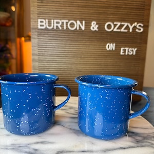May include: Two blue mugs with a white speckled pattern and a handle on the side. Behind the mugs is a brown letter board with white text that reads BURTON & OZZY'S ON ETSY. The mugs are positioned on a white surface with grey veining.