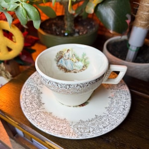 May include: Cream-colored teacup and saucer set. The teacup features a handle and a decorative image of two women in a garden. The saucer and the rim of the teacup are adorned with a gold filigree pattern. The set is placed on a wooden surface.
