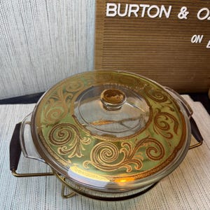 May include: A round glass casserole dish with a clear lid featuring a gold scrollwork pattern. The dish is set in a gold-toned metal wire frame with two dark brown handles on the sides, providing a stable base for the glass container.