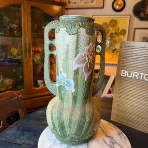 Può includere: Un vaso in ceramica alto con corpo color crema dalla texture punteggiata, decorato con fogliame verde in rilievo e fiori di iris dipinti nelle tonalità del blu e del rosa. Il vaso presenta due manici traforati e un bordo strutturato.