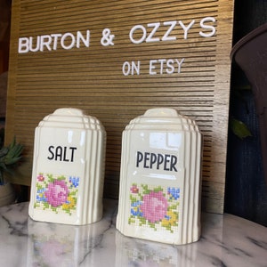 May include: A set of two vintage salt and pepper shakers with a floral design. The shakers are white with pink, blue, and yellow flowers. The words "SALT" and "PEPPER" are printed in black on the front of the shakers.