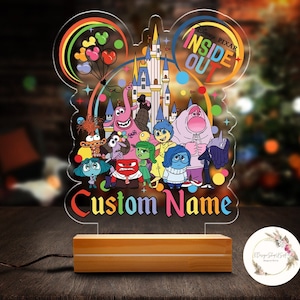 May include: A colorful acrylic night light featuring characters from the Disney Pixar movie Inside Out. The light is shaped like Mickey Mouse ears with a silhouette of Cinderella's castle in the center. The characters are arranged around the castle and the words "Inside Out" are written in a colorful arc above the castle. The light has a wooden base and a cord for plugging it in. The words "Custom Name" are written below the characters.