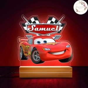 May include: A red acrylic night light featuring Lightning McQueen from the Cars movie. The design includes the name "Samuel" above the car, with checkered flags and a wooden base. The light is illuminated.