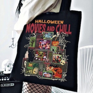 May include: Black tote bag featuring a Halloween-themed design. The design includes the text "HALLOWEEN MOVIES AND CHILL" and various spooky movie scenes. A black cat, spiderwebs, and a jack-o'-lantern are also featured.
