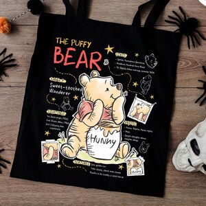 May include: Black tote bag featuring Winnie the Pooh with the text "The Puffy Bear". The design includes illustrations and text about Pooh's characteristics and interests. The bag has black handles and is decorated with Halloween-themed items.