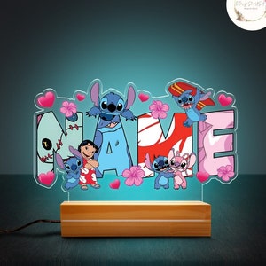 May include: A light-up name sign with the word "NAME" in blue, pink, and red. The letters are decorated with Stitch and Angel characters from the Disney movie Lilo & Stitch. The sign is on a wooden base.