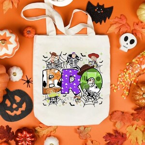 Puede incluir: Una bolsa de lona blanca con un gr&aacute;fico colorido de los personajes de Toy Story, Woody, Buzz Lightyear y Jessie. Los personajes est&aacute;n dispuestos para deletrear la palabra "BRO" con un dise&ntilde;o de telara&ntilde;a en el fondo.