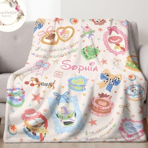 May include: A white fleece blanket with a colorful pattern featuring characters from the Toy Story franchise. The blanket has a pink and white bow, stars, and the name "Sophia" written in pink.