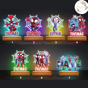 May include: Seven different designs of personalised night lights featuring Spider-Man characters. Each light has a different character and name, including Thomas and Sophia. The lights are on a wooden base and have a colourful glow.