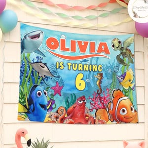 May include: A personalized birthday banner featuring the name "Olivia" and the phrase "is turning 6." The banner showcases colorful illustrations of characters from the movie "Finding Nemo" against a blue underwater background.