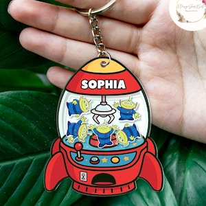 May include: A red, yellow, and white rocket-shaped keychain featuring the name "SOPHIA" and a claw machine design with cartoon aliens. The keychain has a silver keyring and chain.