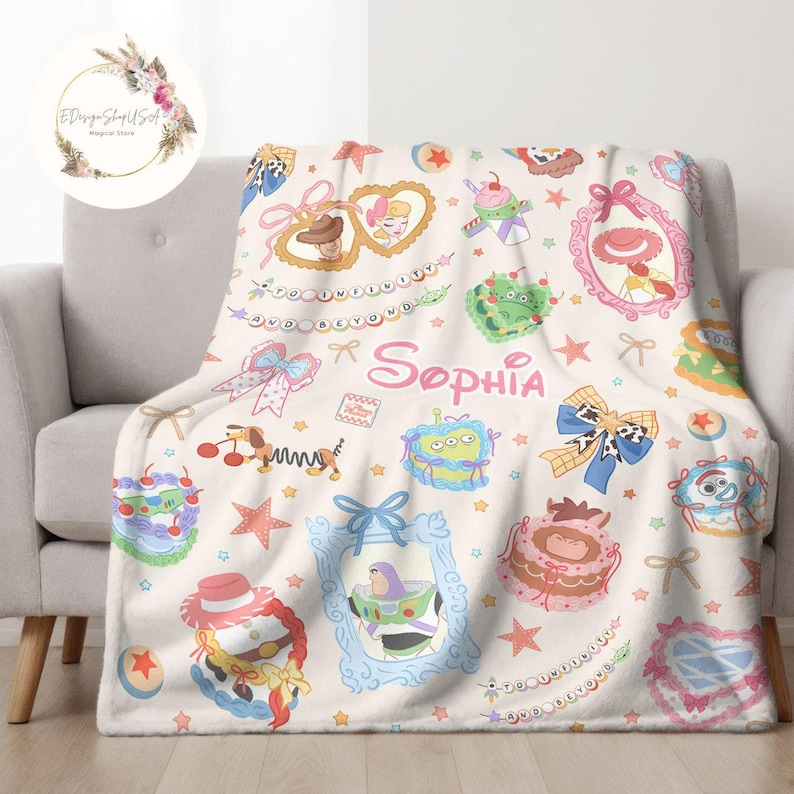 May include: A white fleece blanket with a colorful pattern featuring characters from the Toy Story franchise. The blanket has a pink and white bow, stars, and the name "Sophia" written in pink.