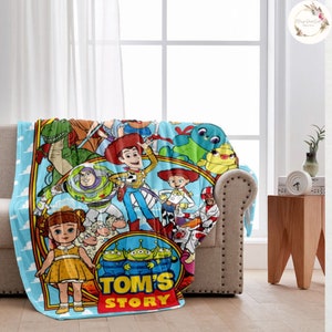 Puede incluir: Una manta de franela azul y roja con una ilustraci&oacute;n colorida de personajes de la franquicia de pel&iacute;culas Toy Story. La manta presenta a Woody, Buzz Lightyear, Jessie, Rex y otros personajes. El texto "TOM'S STORY" est&aacute; impreso en rojo en la manta.