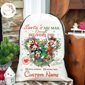May include: A white canvas Santa sack with a drawstring closure. The sack features a Mickey Mouse-shaped design with Mickey, Minnie, Donald Duck, Goofy, and Pluto characters. The text on the sack reads "Santa's Air Mail Overnight Delivery For" and "Custom Name".