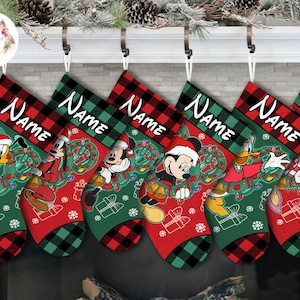 May include: Five personalized Christmas stockings with a red and green plaid pattern. Each stocking features a different Disney character: Pluto, Goofy, Mickey Mouse, Donald Duck, and Daisy Duck. The stockings are decorated with a green wreath, red and white lights, and a gift box.