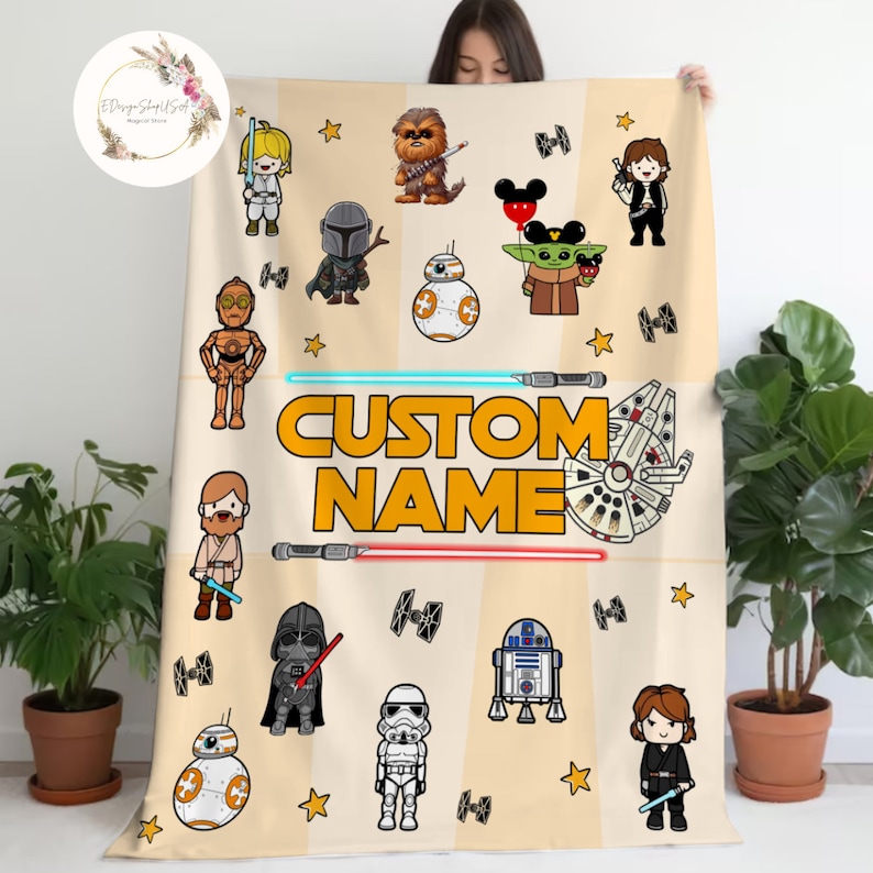 May include: A beige fleece blanket with a custom name in yellow text. The blanket features a variety of cartoon characters from Star Wars and Disney, including Yoda, Darth Vader, R2-D2, and Mickey Mouse. The blanket is decorated with yellow stars and red and blue lightsabers.