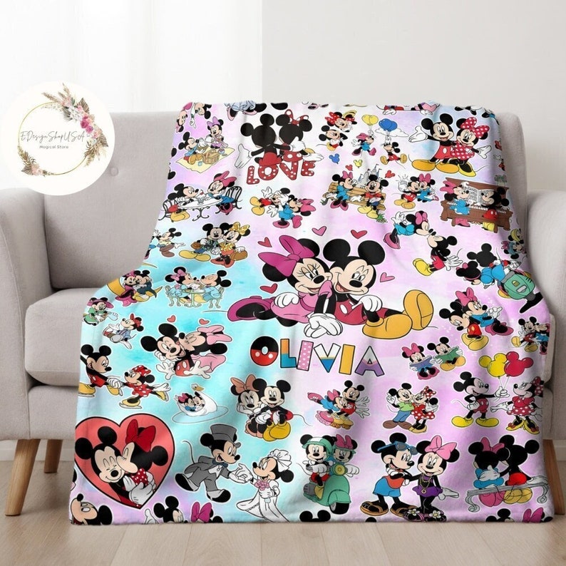 May include: A colourful fleece blanket featuring a repeating pattern of Mickey Mouse and Minnie Mouse in various poses. The blanket has a light blue and pink background with hearts and the name "Olivia" printed in rainbow letters.